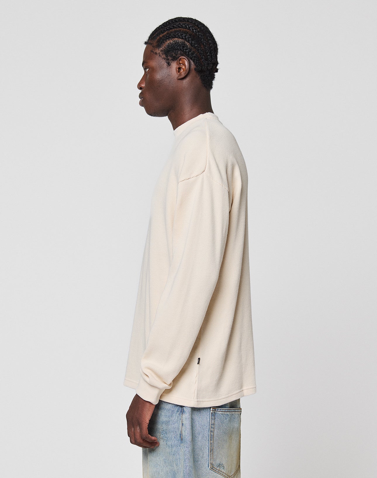 A person with braided hair stands in profile against a light gray background wearing the LIVE FAST DIE YOUNG Blank Waffle Longsleeve in cream, an oversize 100% Baumwolle shirt with a subtle flag-label detail, paired with light blue jeans.