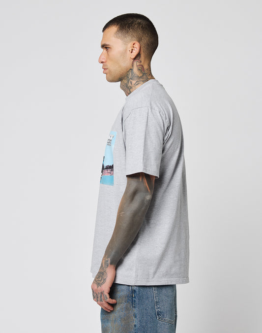 Man with tattoos stands in profile wearing the LIVE FAST DIE YOUNG LFDY Crash Kid Tee, a light gray graphic t-shirt in Regular Fit, paired with blue jeans against a plain light background.