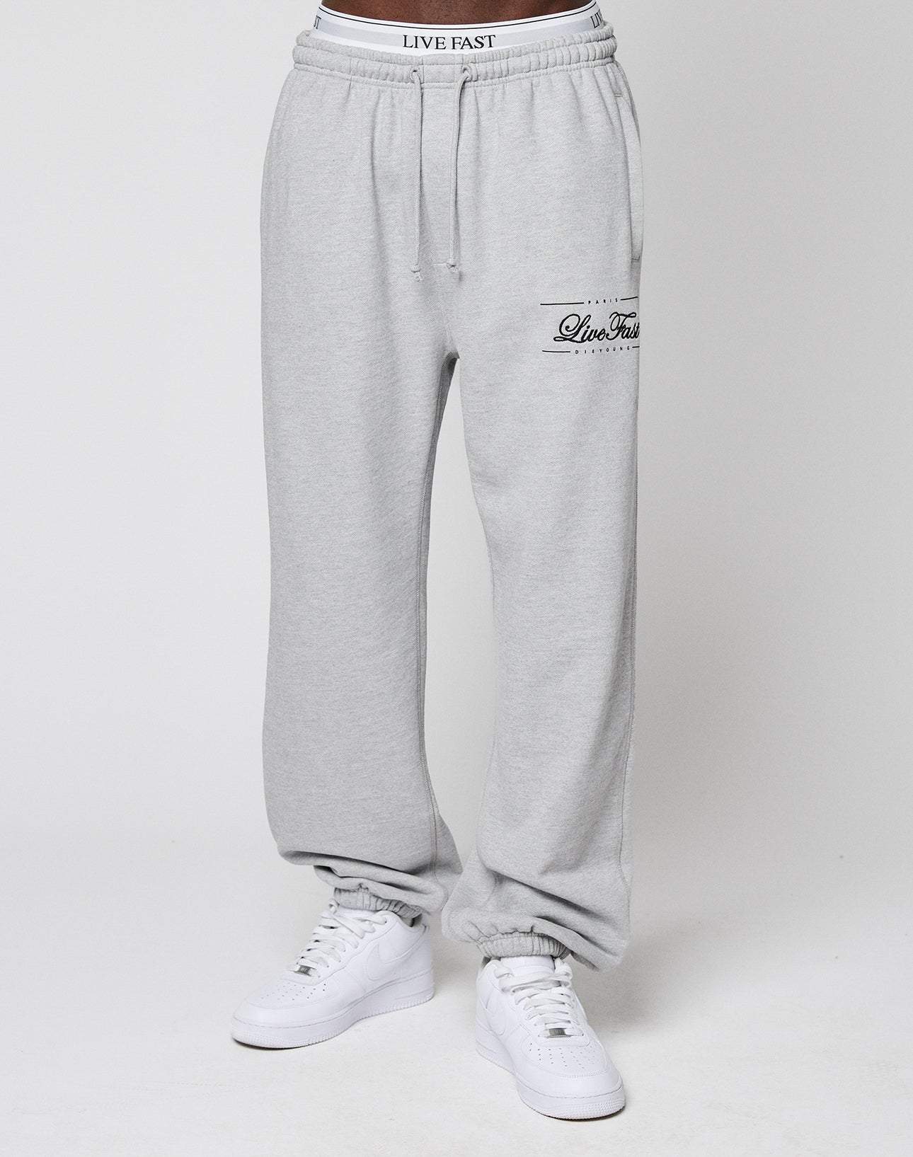 Someone wears LIVE FAST DIE YOUNG Wide Cuff Jogger sweatpants in light gray with "Live Fast" on the left thigh, wide fit, and elastic waistband, paired with white sneakers against a plain white background.