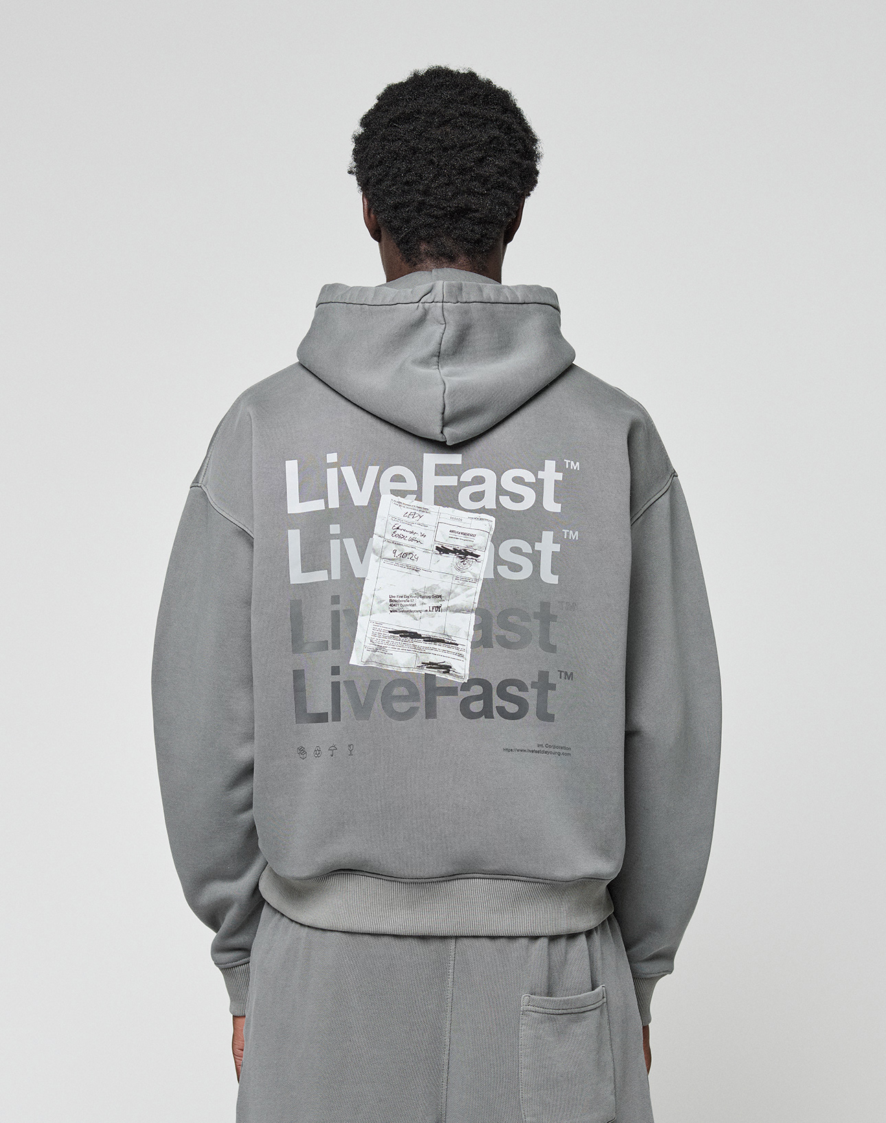 A person wears the Basic Evidence Hooded by LIVE FAST DIE YOUNG, an oversize gray hoodie with “Live Fast” repeated in white and a crumpled paper graphic on the back, shown from behind.