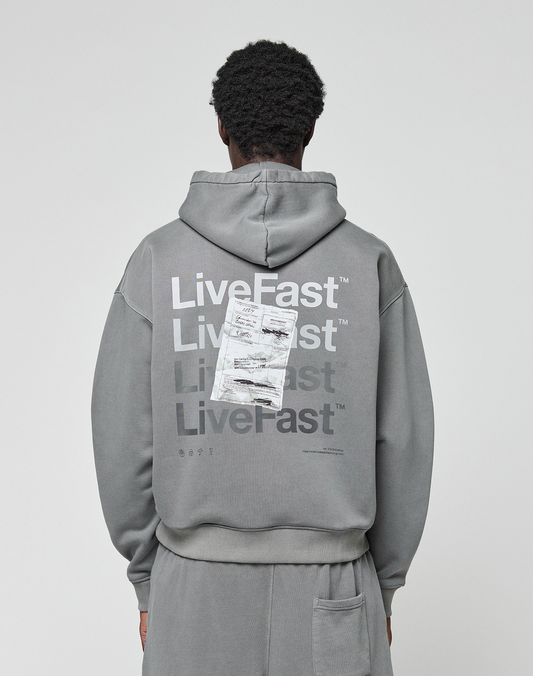 A person wears the Basic Evidence Hooded by LIVE FAST DIE YOUNG, an oversize gray hoodie with “Live Fast” repeated in white and a crumpled paper graphic on the back, shown from behind.