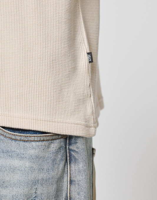 Close-up of a person wearing light blue jeans and the LIVE FAST DIE YOUNG Blank Waffle Longsleeve, an oversize textured beige top with a small black flag label near the hem. The background is plain and light-colored.
