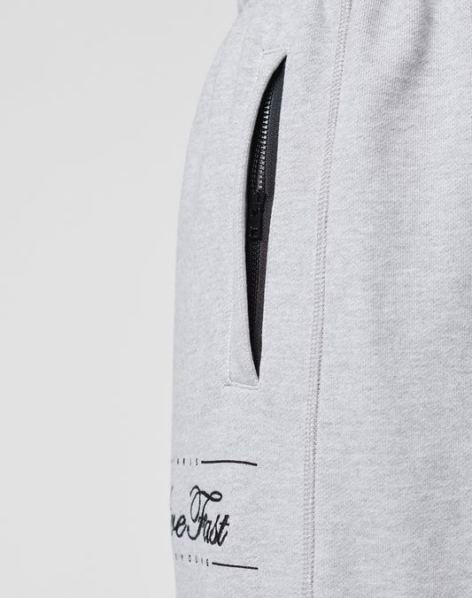 Close-up of the LIVE FAST DIE YOUNG Wide Cuff Jogger, a wide fit, light grey French Terry sweatpant with a black zippered pocket and partial black text near the pocket, shown on a plain background.