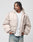 A man with braided hair wears the LIVE FAST DIE YOUNG LFDY Winter Bomber Jacket in light beige, styled oversize over a white T-shirt and blue jeans, standing with hands in pockets and looking left against a plain light background.