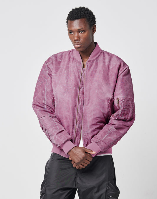 A person wears the LIVE FAST DIE YOUNG Washed Bomber Jacket in mauve with black cargo pants, standing against a plain light gray background and looking slightly to the side with hands clasped in front.