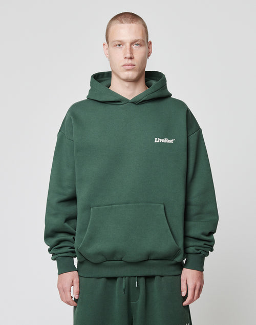 A person with short hair wears the LIVE FAST DIE YOUNG Basic 360 Hooded in dark green and matching sweatpants, standing against a plain white background. The hoodie features LiveFast in small white letters on the left chest.