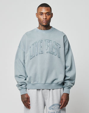 A person wears a light blue Ripped College Sweater by LIVE FAST DIE YOUNG, featuring raised LIVE FAST text on the front, paired with light gray sweatpants, standing against a plain light background.