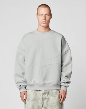 A person with short hair wears the LIVE FAST DIE YOUNG LFDY Wavy Sweater in light gray Oversize Fit, showcasing curved seam details and small white chest text, paired with light gray-green pants against a plain light background.