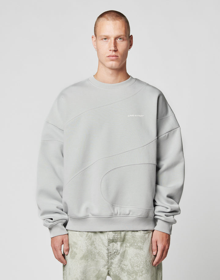 A person with short hair wears the LIVE FAST DIE YOUNG LFDY Wavy Sweater in light gray Oversize Fit, showcasing curved seam details and small white chest text, paired with light gray-green pants against a plain light background.