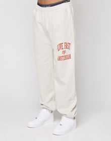 A person wears the LIVE FAST DIE YOUNG Live Fast Amsterdam Loose Jogger in off-white with an elastic waistband and red LIVE FAST AMSTERDAM on the left thigh, made from soft cotton, paired with white sneakers. Only their lower body is visible. | model