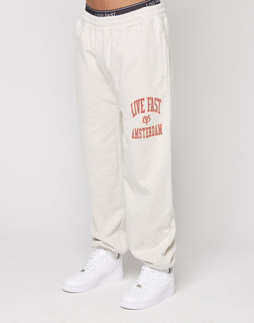 A person wears the LIVE FAST DIE YOUNG Live Fast Amsterdam Loose Jogger in off-white with an elastic waistband and red LIVE FAST AMSTERDAM on the left thigh, made from soft cotton, paired with white sneakers. Only their lower body is visible.