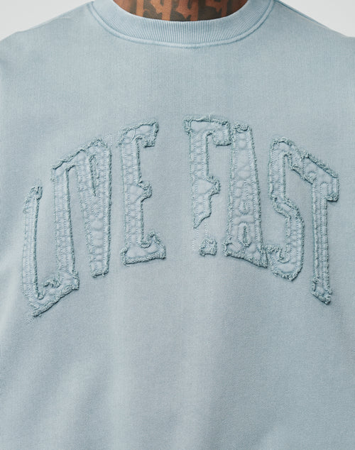 A person wears the Ripped College Sweater by LIVE FAST DIE YOUNG, a light blue sweatshirt with vintage wash and large, distressed “LIVE FAST” applique text arched across the chest. Their tattooed neck is partially visible above the collar.