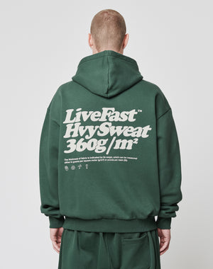 A short-haired person, facing away from the camera, wears a dark green Basic 360 Hooded by LIVE FAST DIE YOUNG in an oversized fit. The Baumwolle-Polyester hoodie features LiveFast HvySweat 360g/m² and smaller text with symbols on the back in white.