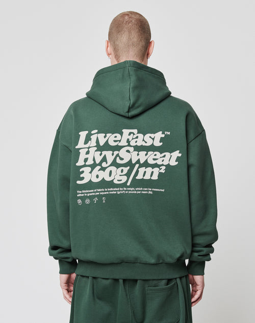 A short-haired person, facing away from the camera, wears a dark green Basic 360 Hooded by LIVE FAST DIE YOUNG in an oversized fit. The Baumwolle-Polyester hoodie features LiveFast HvySweat 360g/m² and smaller text with symbols on the back in white.