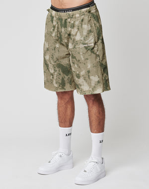 Someone stands against a plain light background wearing LIVE FAST DIE YOUNG Live Fast Nylon Shorts in green and beige camo with lateral pockets and silicone LIVE FAST prints, paired with white crew socks and white sneakers.