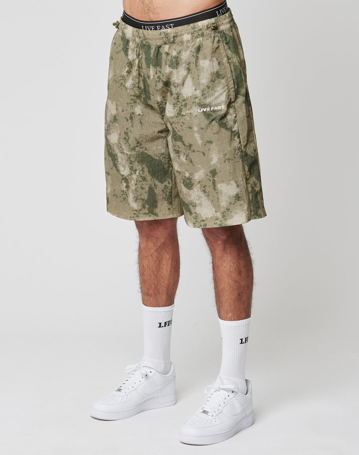 Someone stands against a plain light background wearing LIVE FAST DIE YOUNG Live Fast Nylon Shorts in green and beige camo with lateral pockets and silicone LIVE FAST prints, paired with white crew socks and white sneakers.