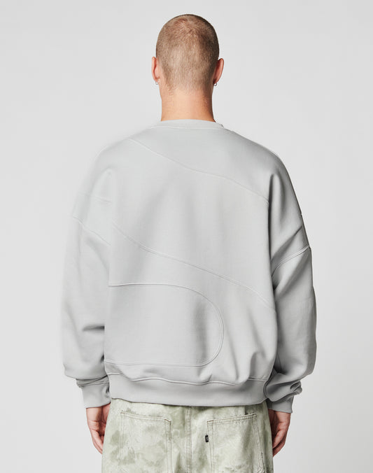 A person with a shaved head is seen from behind, wearing the LIVE FAST DIE YOUNG LFDY Wavy Sweater in light gray, featuring subtle curved stitching and an oversized fit, paired with textured light gray pants. The background is plain and light.