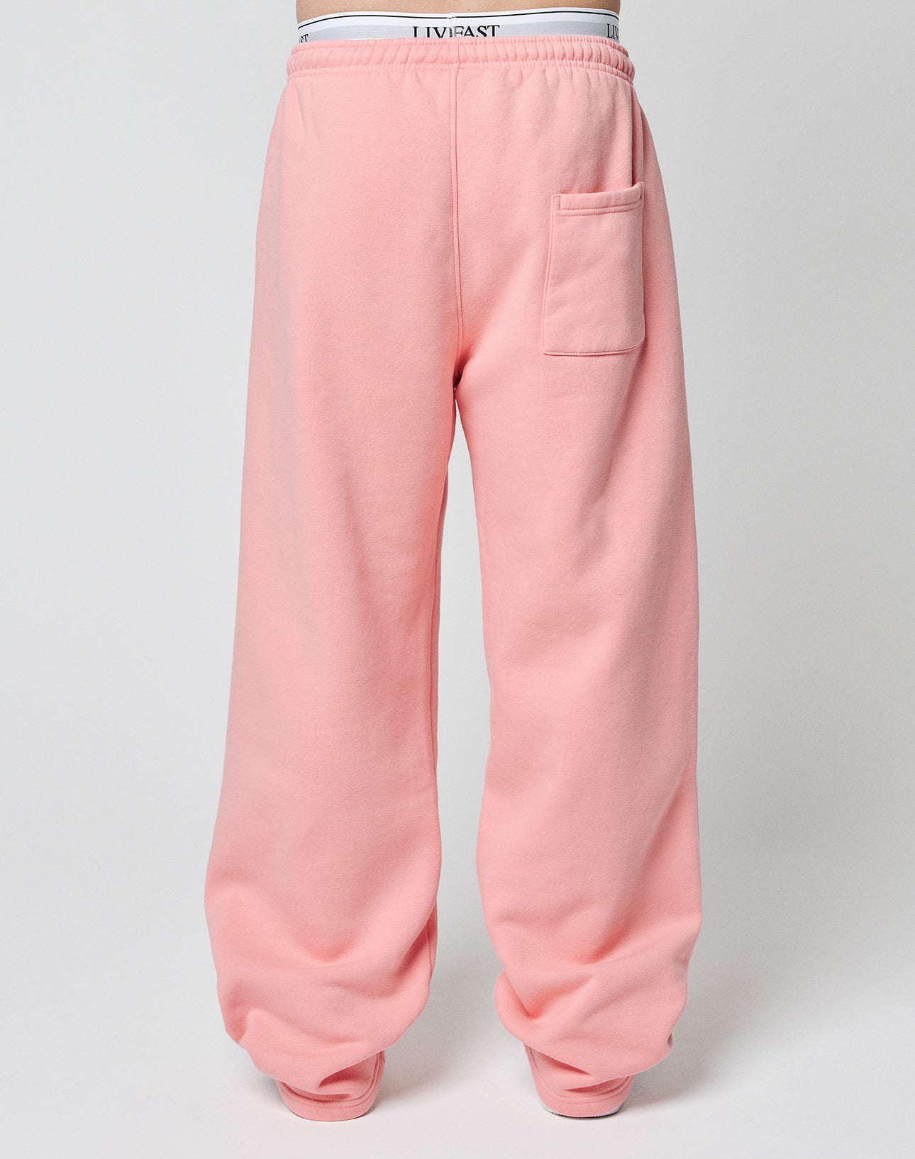 A person wears the LIVE FAST DIE YOUNG Basic Wide Jogger—loose, pink sweatpants with an oversize fit, single back pocket, and bunched ankles—against a plain light background. White underwear waistband shows above the pants.