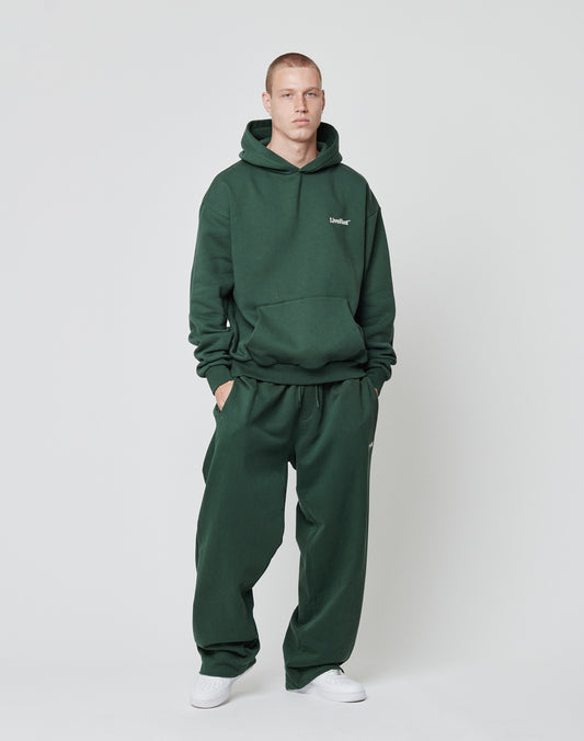A person stands against a plain background wearing LIVE FAST DIE YOUNGs Basic Wide Jogger in dark green with a matching hoodie and an elastic waistband, paired with white sneakers, hand in pocket and a neutral expression.