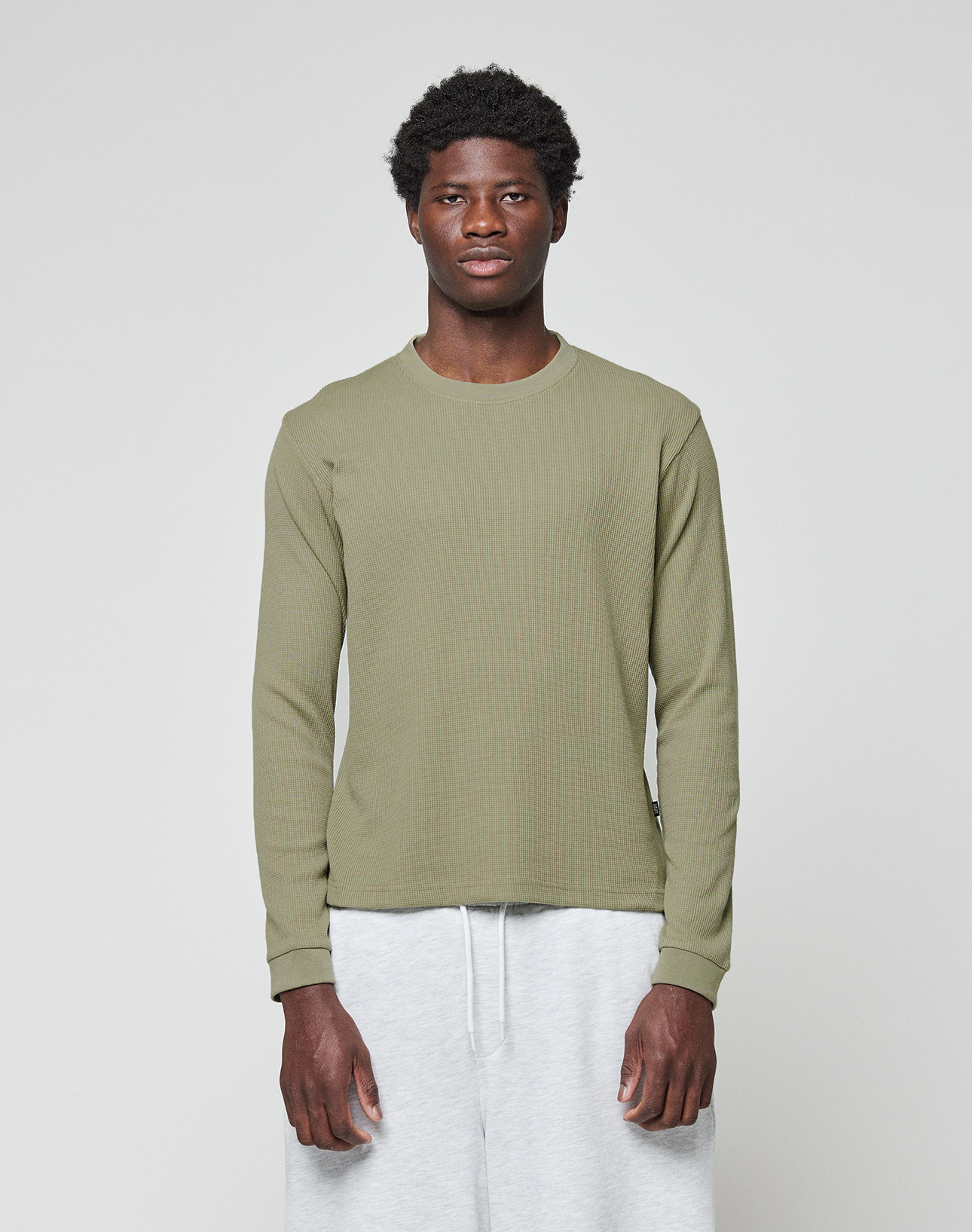A young man faces forward against a plain light gray background, wearing the LIVE FAST DIE YOUNG Basic Waffle Longsleeve in olive green and light gray sweatpants.