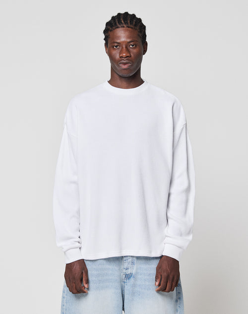 A man with braided hair wears the LIVE FAST DIE YOUNG Blank Waffle Longsleeve in white and light blue Oversize Fit jeans, standing against a plain light gray background, facing the camera with a neutral expression.