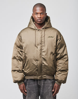A person faces forward in a shiny olive green LFDY Winter Bomber Jacket by LIVE FAST DIE YOUNG, featuring an oversize fit, hood with drawstrings, chest logo embroidery, and paired with dark jeans against a plain light background.