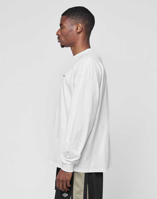A man with short hair stands in profile against a light background, wearing a LIVE FAST DIE YOUNG Basic Longsleeve in white and black shorts with beige stripes.