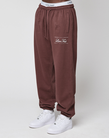 A person stands against a plain white background wearing LIVE FAST DIE YOUNG Wide Cuff Jogger in maroon with a white elastic waistband and white sneakers.