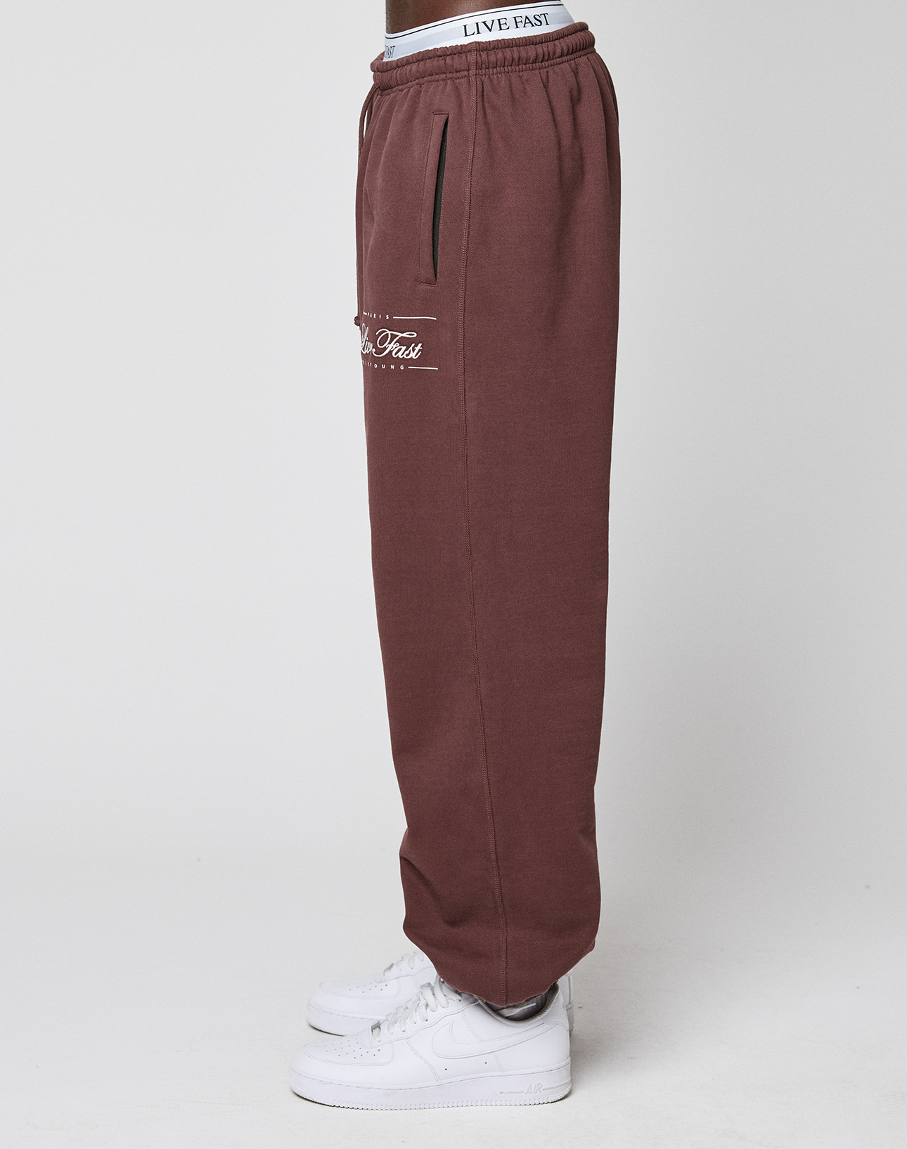 A person stands sideways against a white background wearing maroon Wide Cuff Jogger by LIVE FAST DIE YOUNG, featuring French Terry, a white elastic waistband with LIVE FAST text, and white sneakers. "LIVE FAST" appears on the pocket.