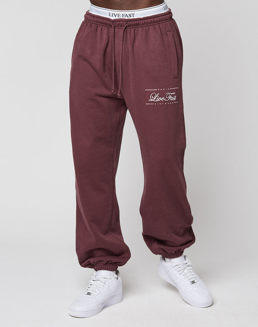 A person in maroon Wide Cuff Jogger sweatpants by LIVE FAST DIE YOUNG, featuring "Live Fast" text on the left thigh and a branded waistband, paired with white sneakers against a plain neutral background.