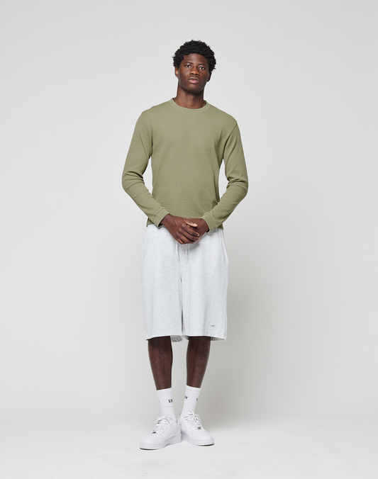A man stands against a white background, facing forward with hands clasped, wearing LIVE FAST DIE YOUNG Basic Shorts—light gray, knee-length with elastic waistband—an olive green long-sleeve shirt, white socks, and sneakers.