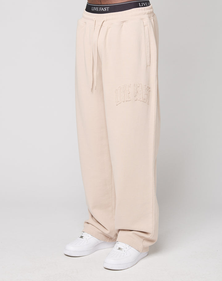 A person in beige Ripped College Wide Jogger sweatpants from LIVE FAST DIE YOUNG, featuring LIVE FAST embroidery on the left thigh and a visible black waistband, paired with white sneakers, stands against a white background. | model