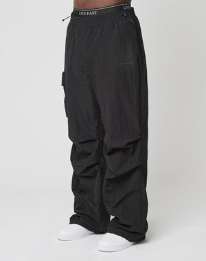 Person wearing Live Fast Nylon Pants by LIVE FAST DIE YOUNG—loose black cargo pants with elastic waistband and cargo pockets, featuring LIVE FAST text, paired with white sneakers against a plain white background.