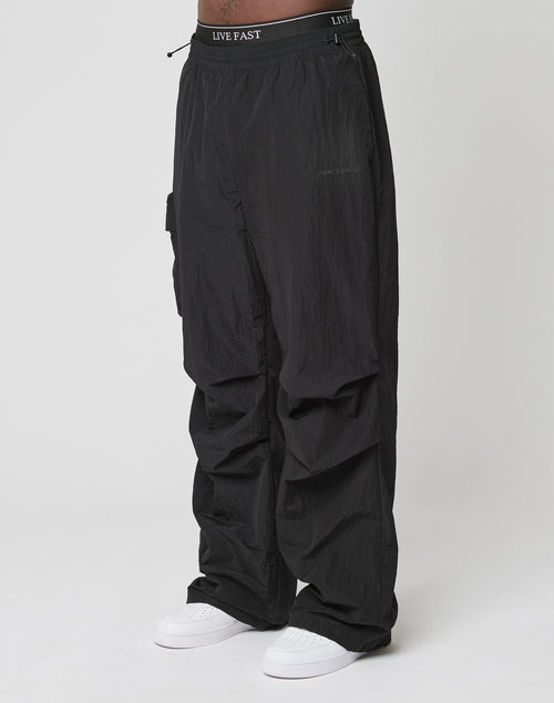 Person wearing Live Fast Nylon Pants by LIVE FAST DIE YOUNG—loose black cargo pants with elastic waistband and cargo pockets, featuring LIVE FAST text, paired with white sneakers against a plain white background.