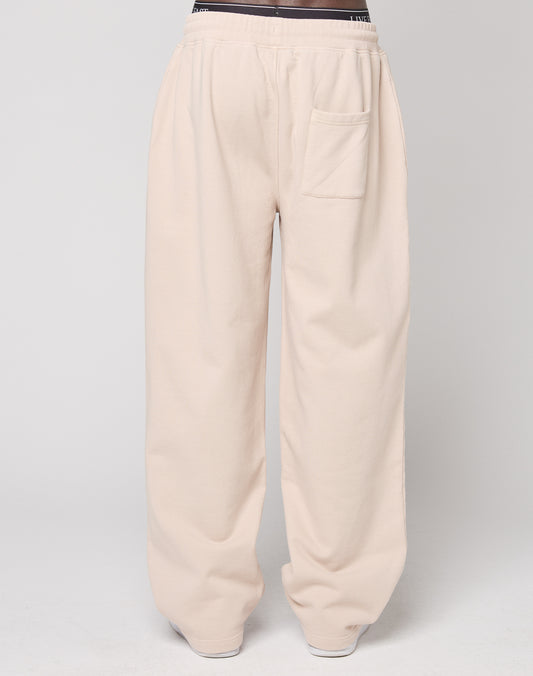 A person wears LIVE FAST DIE YOUNGs Ripped College Wide Jogger, wide fit sweatpants in light beige Baumwolle with an elastic waistband and single front pocket, standing against a plain light gray background.