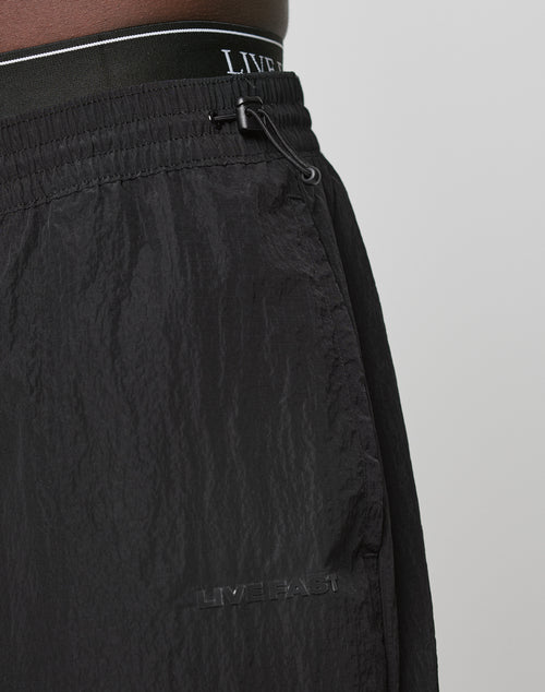 Close-up of a person wearing Live Fast Nylon Pants by LIVE FAST DIE YOUNG, featuring black textured fabric, an elastic waistband with drawstring, embossed LIVEFAST branding on the Cargotasche and waistband. Plain light gray background.