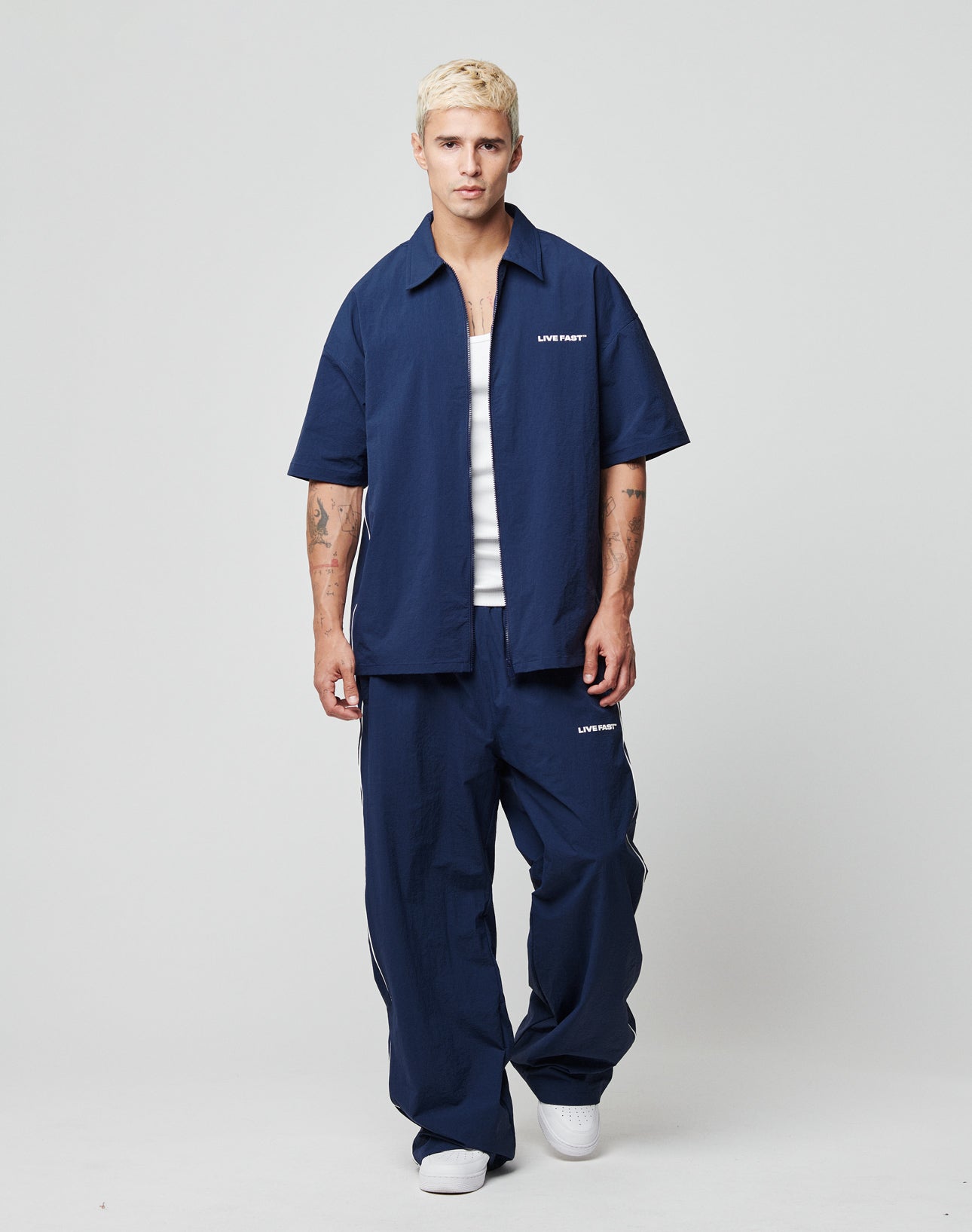 A person with short blond hair wears navy blue LFDY Ripstop Trackpants and a matching shirt from LIVE FAST DIE YOUNG, both with silicone print logos. They pair this with a white tank top and sneakers, standing against a plain light background.