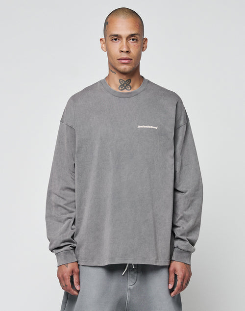 A person with a shaved head and neck tattoo models the LIVE FAST DIE YOUNG Basic Longsleeve—an oversize fit, 100% cotton gray shirt with a subtle chest logo—paired with gray pants against a plain white background.