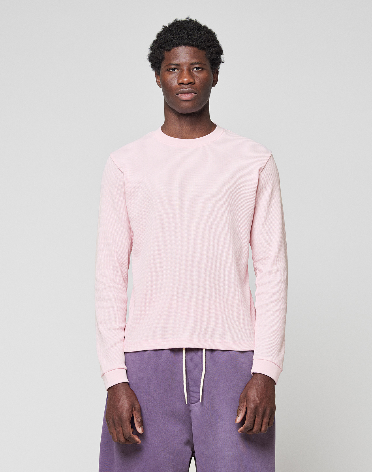 A man stands facing forward, wearing a LIVE FAST DIE YOUNG Basic Waffle Longsleeve in light pink and loose-fitting purple Oversize Fit pants with a white drawstring, set against a plain light gray background.