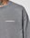 A close-up of a person wearing the LIVE FAST DIE YOUNG Basic Longsleeve, an oversize fit gray sweatshirt with a white LiveFastDieYoung chest logo. Part of a floral neck tattoo is visible above the collar. Made from 100% Baumwolle.