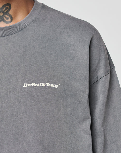 A close-up of a person wearing the LIVE FAST DIE YOUNG Basic Longsleeve, an oversize fit gray sweatshirt with a white LiveFastDieYoung chest logo. Part of a floral neck tattoo is visible above the collar. Made from 100% Baumwolle.
