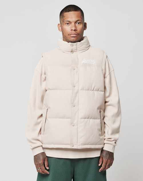 A man with short hair and hand tattoos wears the LIVE FAST DIE YOUNG Basic Vest in beige over a beige sweatshirt and green shorts, standing against a plain light background and looking directly at the camera.