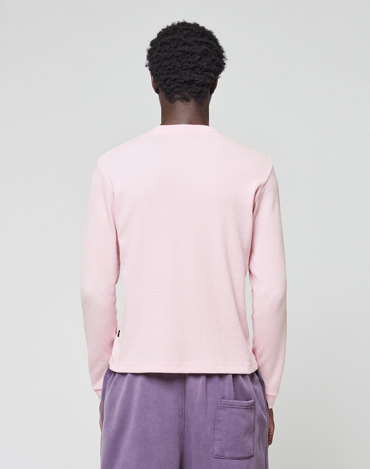 A person with short curly hair, seen from behind, wears a light pink LIVE FAST DIE YOUNG Basic Waffle Longsleeve with Ripp-Bündchen and purple shorts with a back pocket, made of soft cotton, against a plain light gray background.