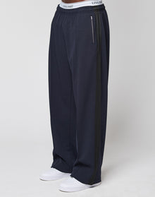 Wearing the LFDY Striped Jogger by LIVE FAST DIE YOUNG, featuring a loose fit, navy blue color, dark Fischgräten-Tape side stripe, and a white LIVE FAST waistband, paired with white sneakers against a plain light background. | model