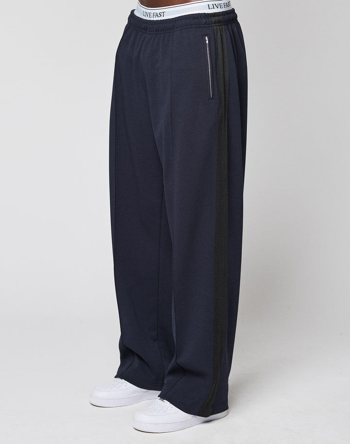 Wearing the LFDY Striped Jogger by LIVE FAST DIE YOUNG, featuring a loose fit, navy blue color, dark Fischgräten-Tape side stripe, and a white LIVE FAST waistband, paired with white sneakers against a plain light background. | model