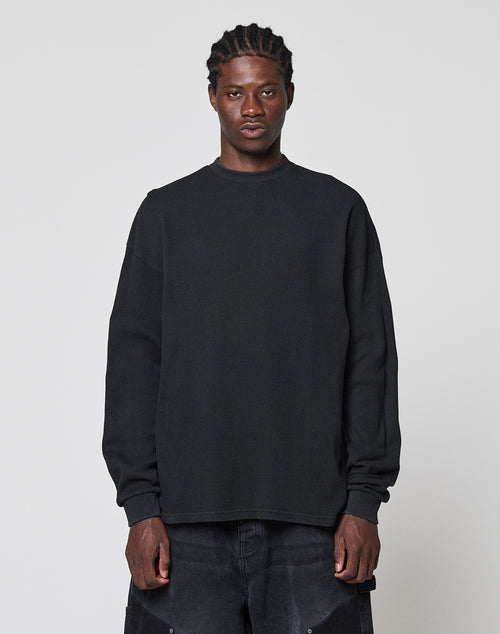 A person with braided hair stands against a plain light gray background, wearing a LIVE FAST DIE YOUNG Blank Waffle Longsleeve in black and black pants, looking directly at the camera with a neutral expression.