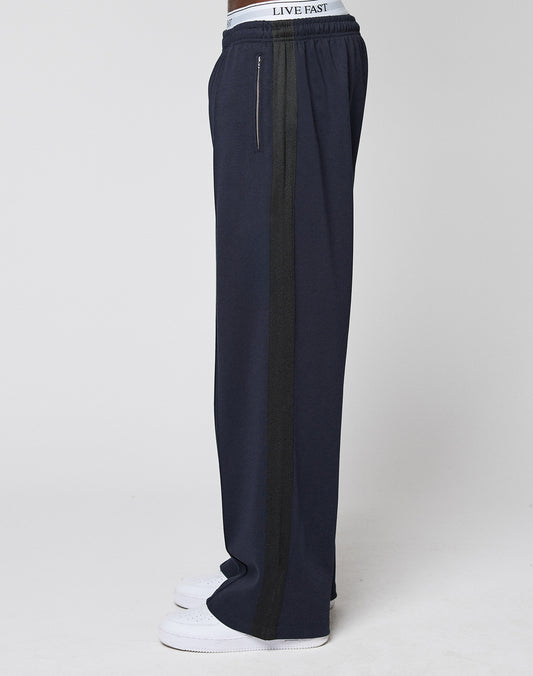 A person in LIVE FAST DIE YOUNG’s LFDY Striped Jogger, featuring a navy blue wide fit, black Fischgräten-Tape side stripe, and an elastic waistband reading LIVE FAST, paired with white sneakers, shown in profile on a plain white background.