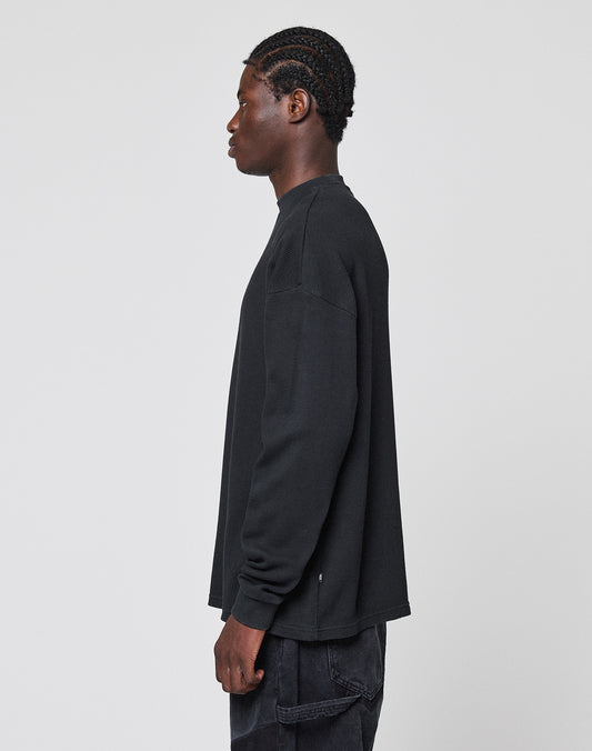 A person with braided hair stands sideways, facing left, wearing the LIVE FAST DIE YOUNG Blank Waffle Longsleeve in black and black pants with Ripp-Bündchen against a plain light gray background.