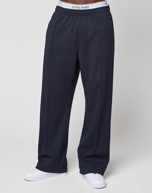 A person wearing loose-fitting dark navy LFDY Striped Jogger sweatpants by LIVE FAST DIE YOUNG, paired with white sneakers and a visible white LIVE FAST waistband, stands against a plain light background.
