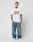 A tattooed man faces the camera in a white LIVE FAST DIE YOUNG LFDY Sixers Tee (Regular Fit, 100% Baumwolle), loose blue jeans, and white sneakers against a gray background. Hands relaxed by his sides. Waschhinweise included.